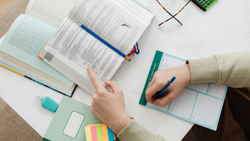 Image of person reading book at desk with computer